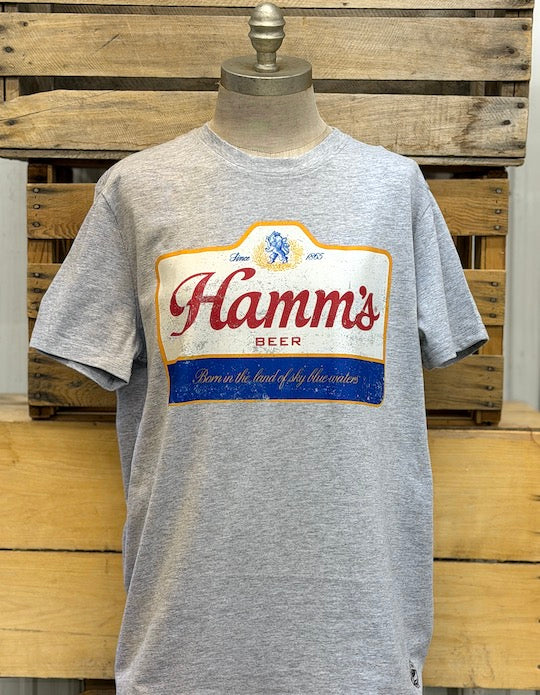 A heather gray short-sleeve t-shirt displayed on a mannequin torso, featuring a large vintage-style 'Hamm's Beer' logo graphic on the chest with red lettering, a cream background, and a blue banner.