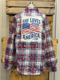 Thumbnail for A long-sleeve plaid button-up shirt displayed on a mannequin torso, featuring a large patriotic back graphic with an American flag and the text 'SHE LOVES JESUS AND AMERICA 100'. The shirt has a red, white, and blue plaid pattern and is photographed against rustic wooden pallets.