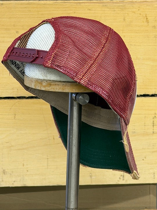 A distressed trucker-style baseball cap with a burgundy mesh front, white mesh back, and a dark green fabric brim, displayed on a wooden stand against a light wood plank background.