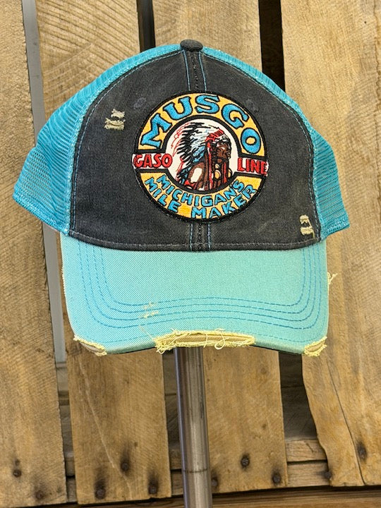 A distressed trucker hat with a dark gray front panel and a light blue brim and mesh back, featuring a circular embroidered Musco Casino Michigan 1969 patch on the front, displayed on a stand against a rustic wooden background.