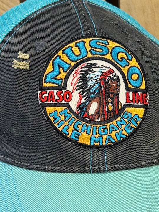 A close-up of a distressed trucker hat featuring a large embroidered circular patch on the front panel. The patch reads 'MUSGO GASO LINE MICHIGAN'S MILE MAKER' with a central Native American graphic.
