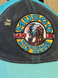 Thumbnail for A close-up of a distressed trucker hat featuring a large embroidered circular patch on the front panel. The patch reads 'MUSGO GASO LINE MICHIGAN'S MILE MAKER' with a central Native American graphic.