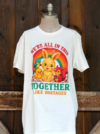 Thumbnail for A white short-sleeve graphic T-shirt displayed on a dress form against a rustic dark wood plank background. The shirt features a colorful Easter-themed graphic with a bunny, chicks, a turtle, a squirrel, and the text 'WE'RE ALL IN THIS TOGETHER LIKE HOSTAGES'.