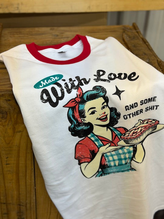 A white ringer t-shirt with a red crew neck, featuring a retro-style graphic of a woman holding a pie and the text 'Made With Love AND SOME OTHER SHIT'. The shirt is laid flat on a rustic wooden table.