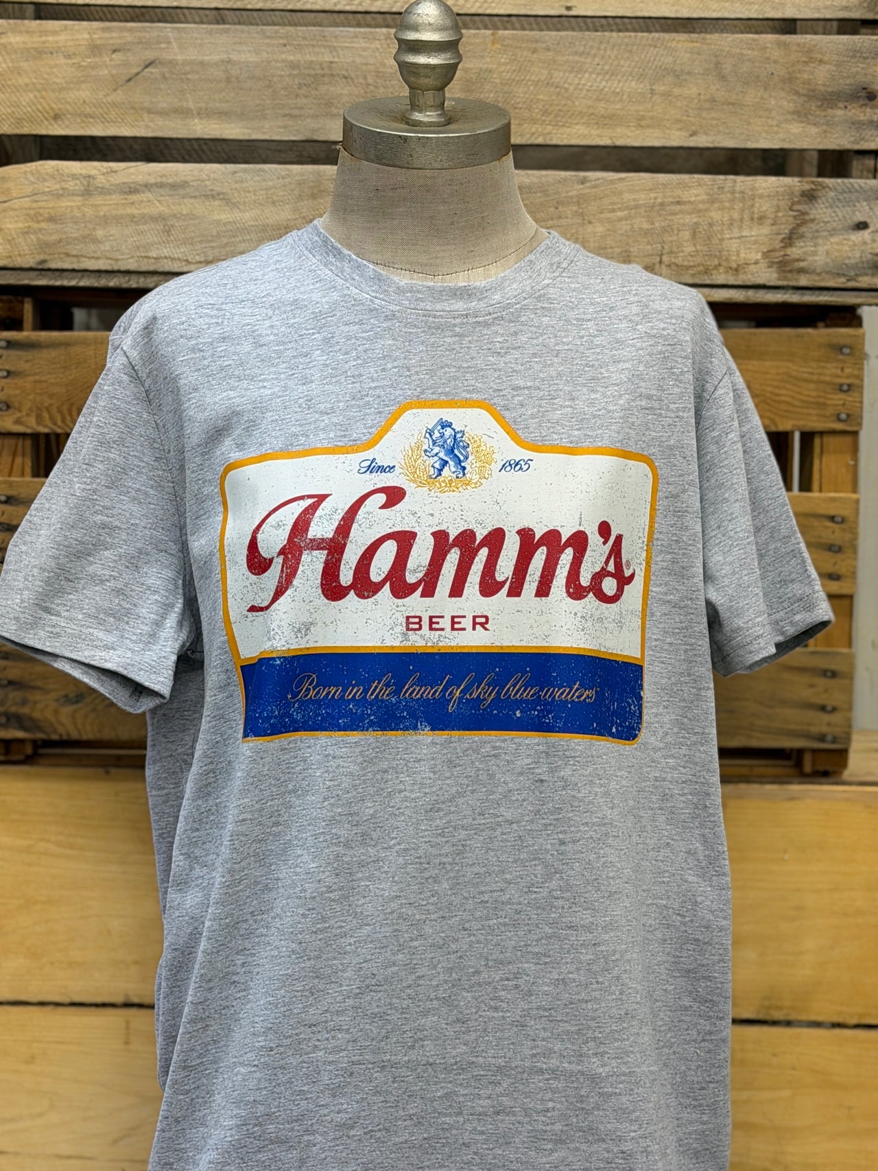 A heather gray short-sleeve t-shirt displayed on a mannequin torso, featuring a large vintage-style printed graphic of the 'Hamm's Beer' logo on the chest