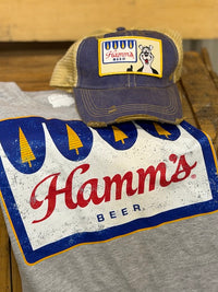 Thumbnail for A gray heather t-shirt featuring a distressed blue, red, and yellow print of the classic Hamm's Beer logo, with a matching navy blue and tan trucker hat resting on top.