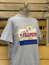 Thumbnail for A heather gray short-sleeve t-shirt displayed on a mannequin torso against a rustic wooden pallet background. The shirt features a large, vintage-style distressed graphic of the Hamm Beer label on the chest.