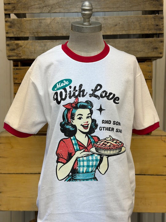 A vintage-style white ringer t-shirt displayed on a mannequin torso against a rustic wooden crate background. The shirt features red ribbed collar and cuffs, and a retro graphic of a pin-up girl holding a pie with the text 'Made With Love AND SON OTHER SHI'.