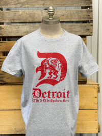 Thumbnail for A heather gray short-sleeve t-shirt displayed on a mannequin torso, featuring a large red distressed graphic of a lion inside a letter D with the word 'Detroit' and smaller text below, set against a rustic wooden crate background.