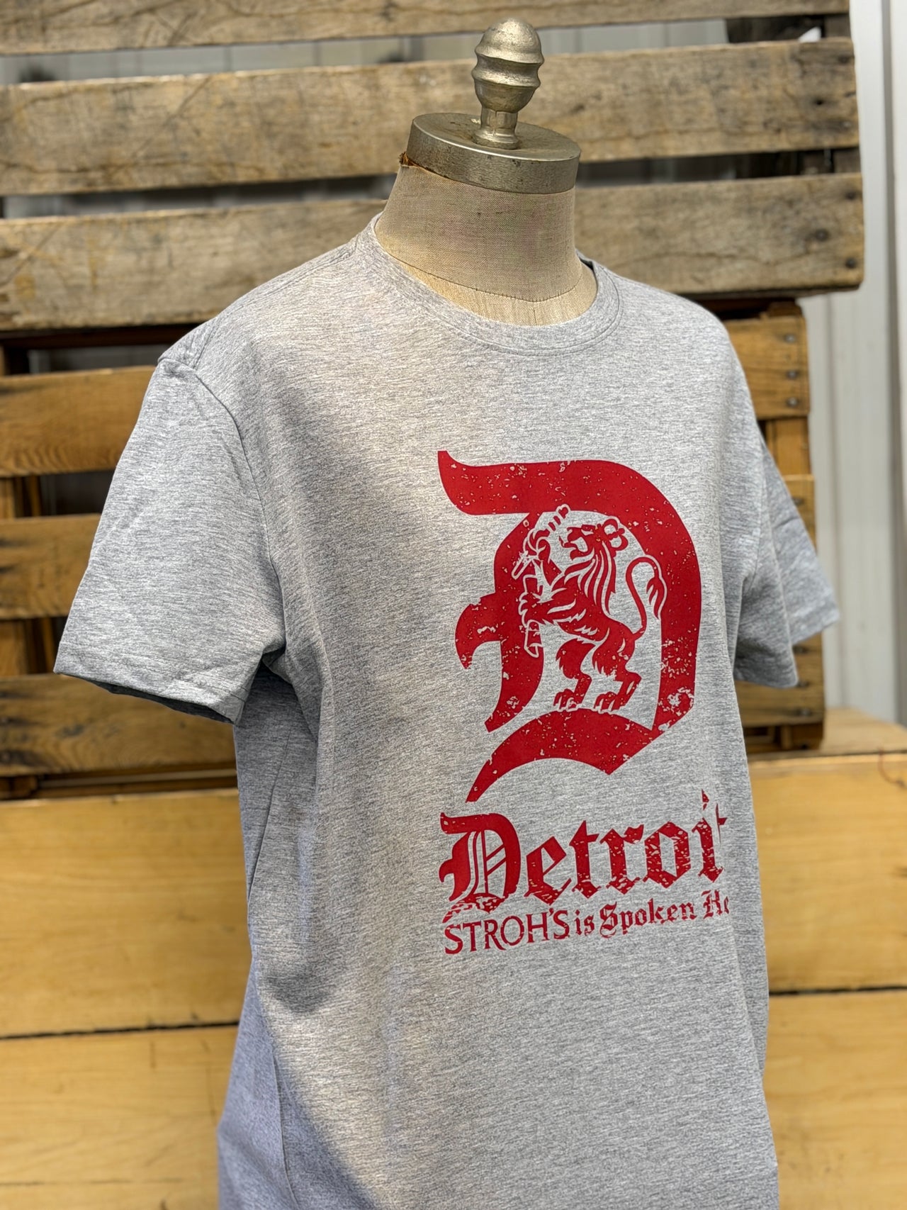 A heather gray short-sleeve t-shirt displayed on a wooden mannequin torso, featuring a distressed red graphic of a lion inside a letter D with the text 'Detroit' and 'STROHS is Spoken Art'. The background consists of rustic wooden pallets.