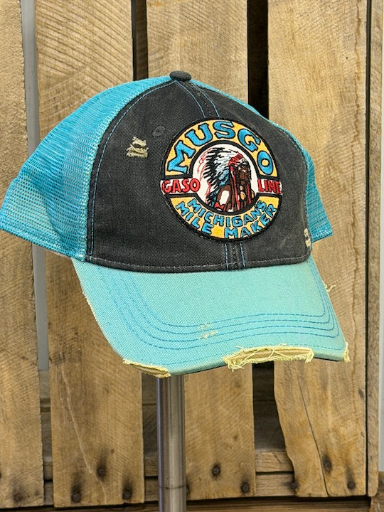 A distressed trucker hat with a light blue brim and mesh back, and a dark charcoal front panel featuring a circular embroidered 'Musco Native American Gasoline' patch. The hat is displayed on a metal stand against a rustic wooden plank background.