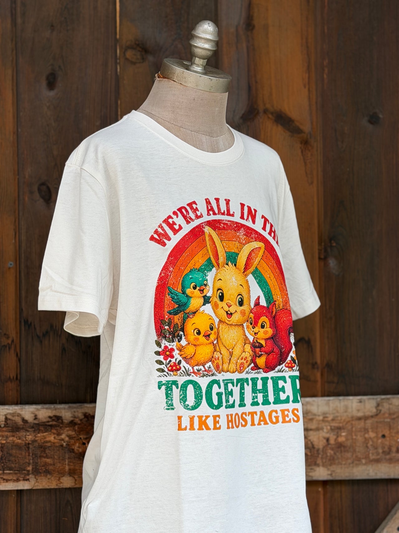 A white graphic t-shirt displayed on a mannequin torso against a rustic dark wood plank background. The graphic features a colorful retro design with a bunny, chicks, a squirrel, and birds under a rainbow, with the text 'WE'RE ALL IN THIS TOGETHER LIKE HOSTAGES'.