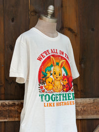 Thumbnail for A white graphic t-shirt displayed on a mannequin torso against a rustic dark wood plank background. The graphic features a colorful retro design with a bunny, chicks, a squirrel, and birds under a rainbow, with the text 'WE'RE ALL IN THIS TOGETHER LIKE HOSTAGES'.