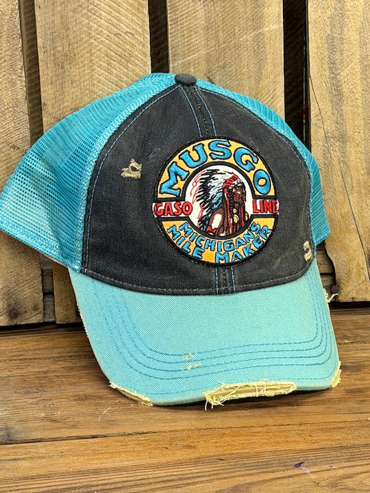 A distressed trucker-style baseball cap with a black front panel and light blue mesh back and brim, featuring a circular embroidered 'Musco Native' patch on the front, set against a rustic wooden background.