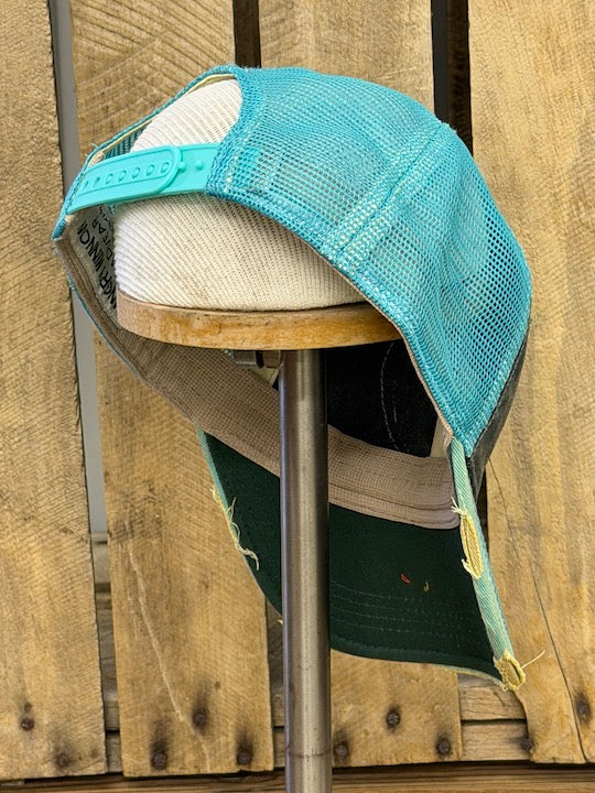 A distressed trucker-style baseball cap with a turquoise blue mesh back, a beige woven front panel, and a dark green brim, displayed on a wooden stand against a rustic wood plank background.