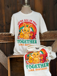 Thumbnail for A white graphic T-shirt displayed on a mannequin torso, with a matching hooded sweatshirt draped over a wooden bench in front. The shirt features a colorful cartoon bunny, chicks, a red squirrel, and the text 'WE'RE ALL IN THIS TOGETHER LIKE HOSTAGES' against a rainbow.