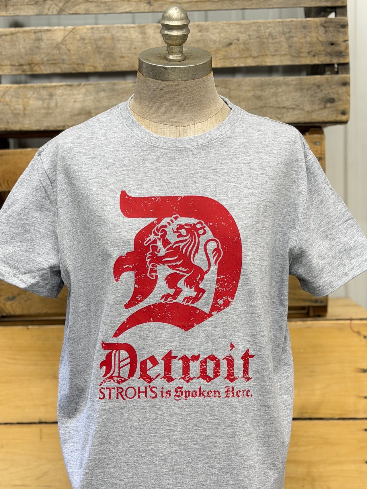 A heather gray short-sleeve t-shirt displayed on a mannequin torso, featuring a large distressed red graphic of a lion inside a letter 'D' with the text 'Detroit' and 'STROHS is Spoken Act.' below it, set against a rustic wooden background.