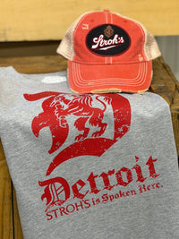 Thumbnail for A heather gray T-shirt with a large distressed red Detroit graphic and text, styled on a wooden surface with an orange-red trucker hat featuring a Stroh's logo on top.